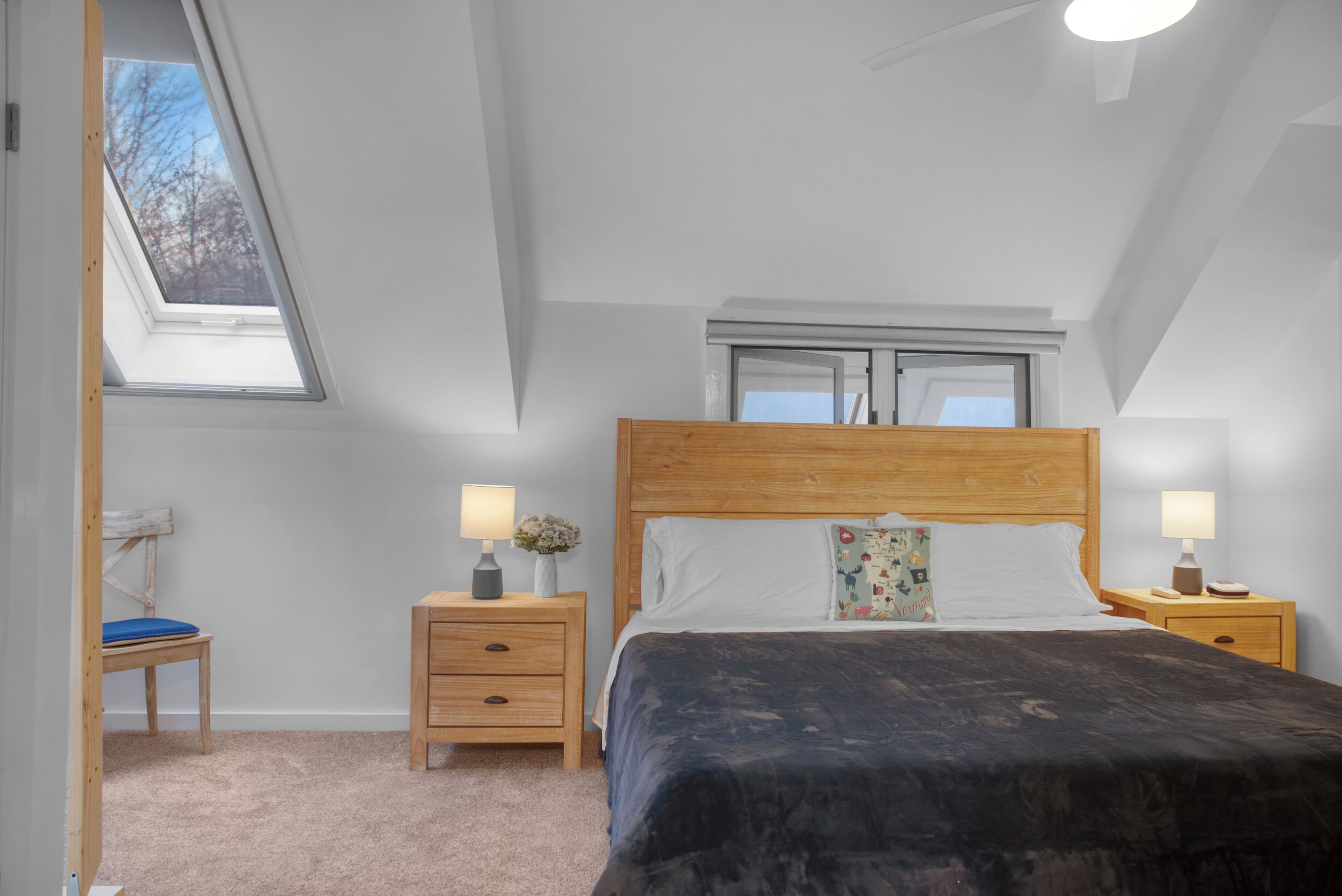 Un dormitorio con tragaluces, cabecera de madera, cama con manta oscura, dos mesitas de noche con lámparas y una silla.