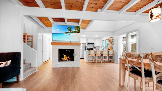 A spacious living area with a fireplace, wooden flooring, a TV mounted on the wall, and a kitchen in the background. - 3 bedroom Chic condo - Community pool and tennis - 1 Mile to Mount Snow (Dover)