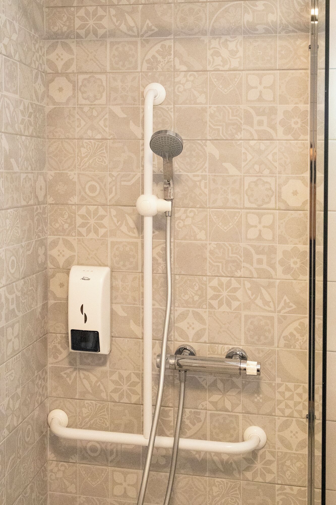 A shower with a handheld showerhead, a wall-mounted soap dispenser, and a handrail.