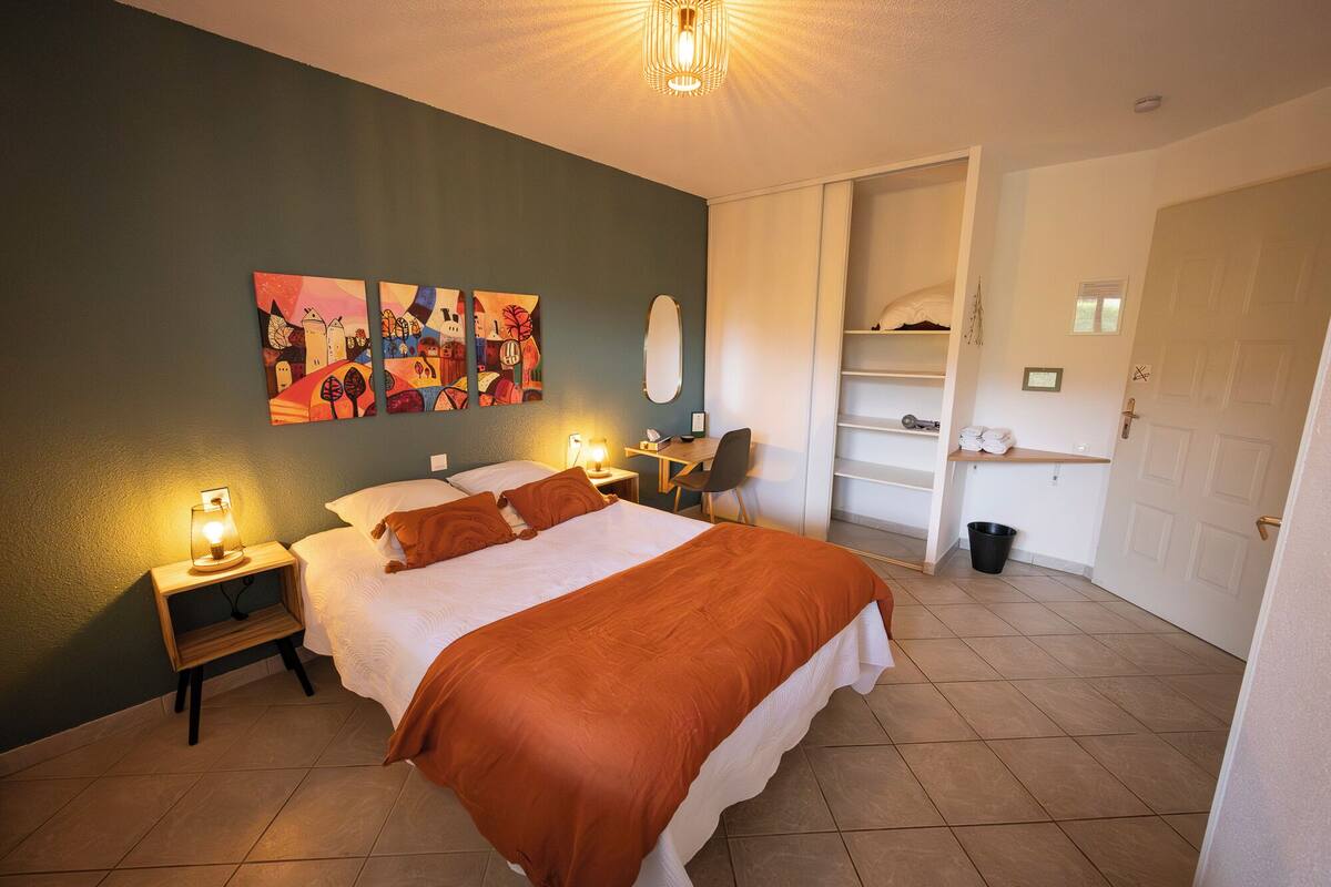 Quarto de hotel com cama, luminárias de cabeceira, escrivaninha, cadeira, armário e banheiro.