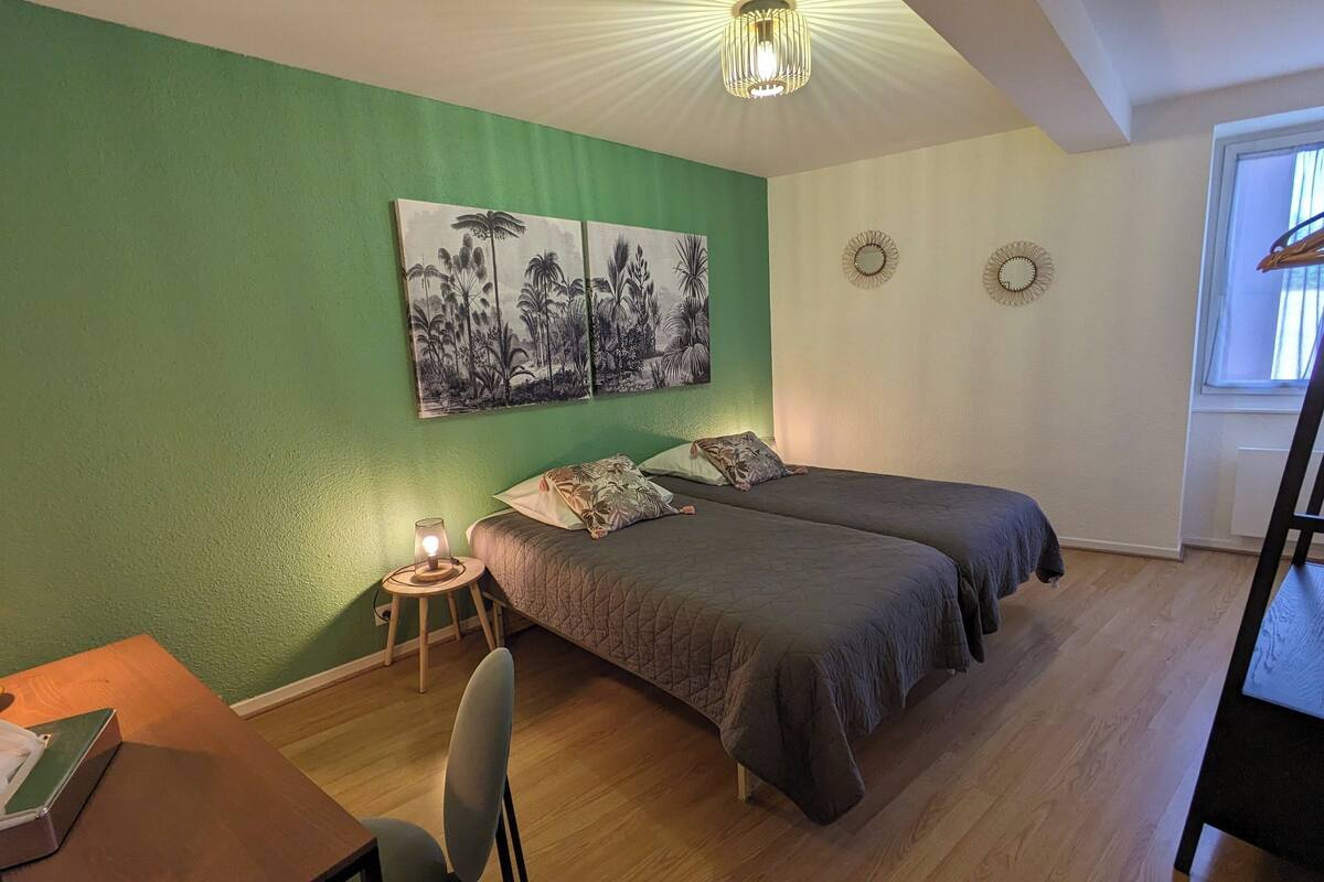 Quarto com duas camas, uma escrivaninha com luminária, uma cadeira, uma mesinha e uma janela.
