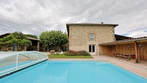 Una piscina con un edificio in pietra sullo sfondo, un patio coperto e un piccolo giardino con piante e un albero.