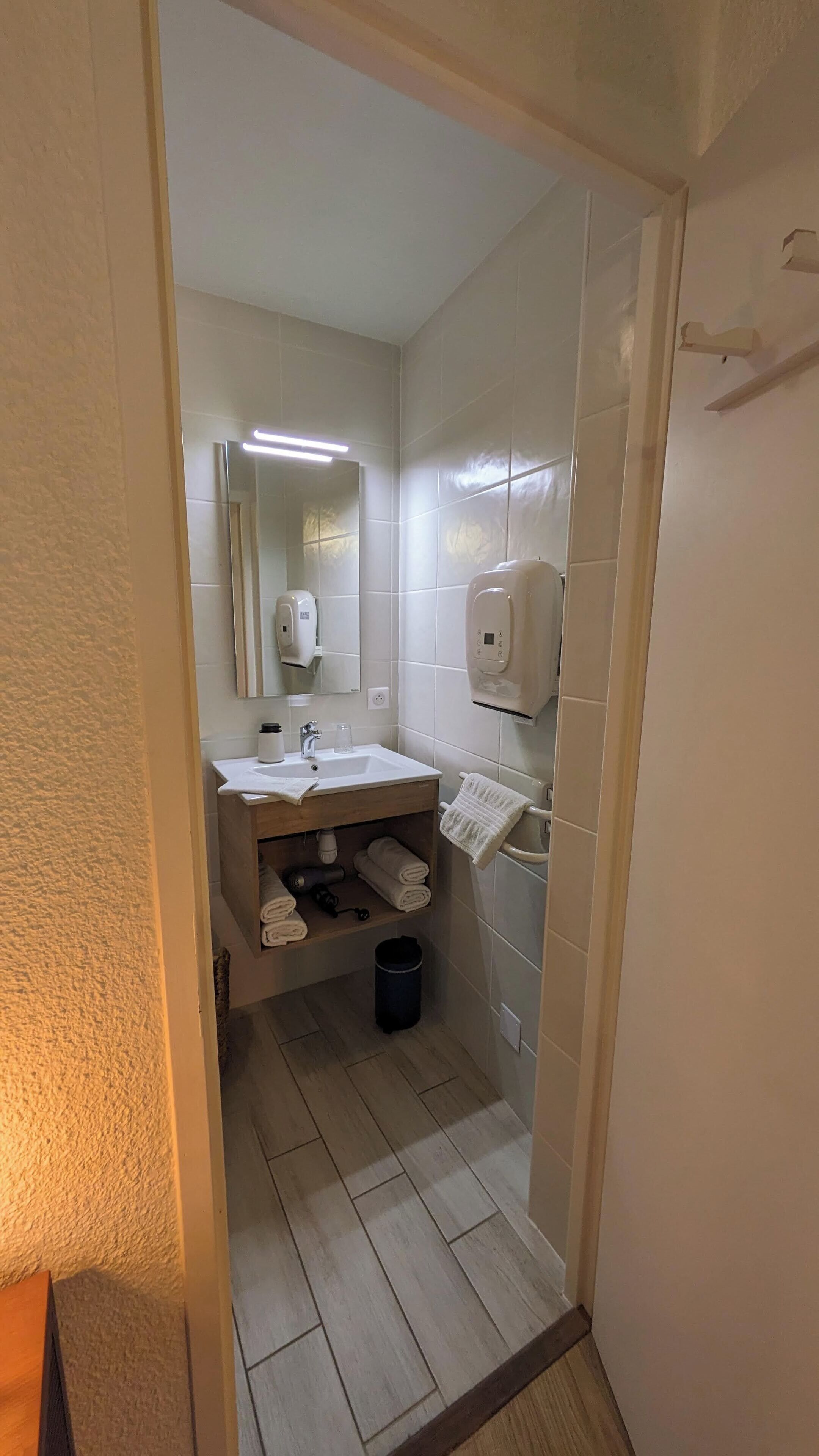 Ein Badezimmer mit einem weißen Waschbecken, einem Spiegel und einem Händetrockner. Ein Handtuchhalter mit Handtüchern und ein Mülleimer sind ebenfalls vorhanden.