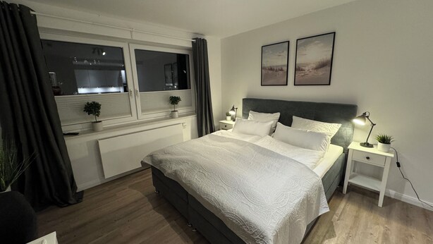 Une chambre moderne avec un grand lit, des tables de chevet avec des lampes, une fenêtre avec des rideaux et deux tableaux encadrés accrochés au mur.