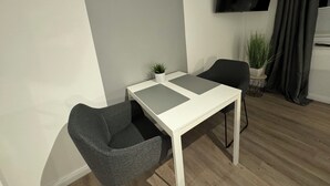 Un salon moderne avec une table blanche, deux fauteuils gris, un téléviseur à écran plat fixé au mur et une plante en pot.