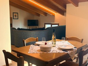 Un coin repas avec une table dressée pour deux personnes, une bouteille de vin blanc et un saladier. Le plafond est en bois et un canapé se trouve au fond de la pièce.