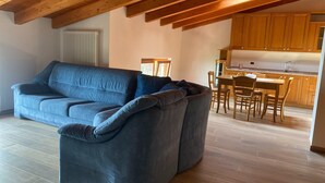 A modern living room with a large grey sofa, wooden ceiling beams, a dining area with a table and chairs, and a kitchen with wooden cabinets.
