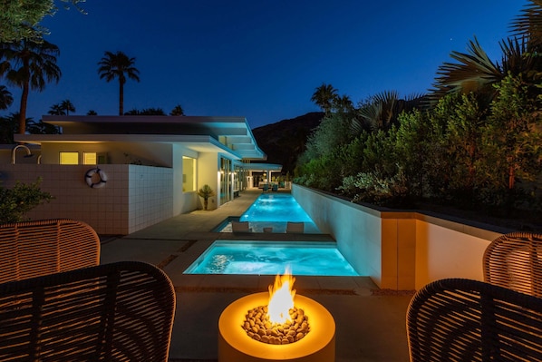 A modern outdoor pool area with a fire feature, surrounded by wicker chairs and palm trees.