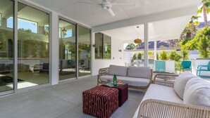 A modern outdoor living area with a ceiling fan, glass doors, a sofa, a coffee table, and a view of greenery and mountains. - Webb House – Mid-Century Modern in Vista Las Palma (Palm Springs)