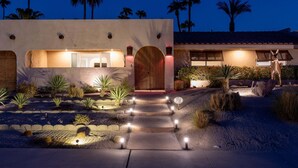 A modern house with a well-lit entrance, a stone pathway, and a desert garden with cacti and palms. - Desert Delight – Palm Springs Hacienda w/ Pool & Spa (Palm Springs)