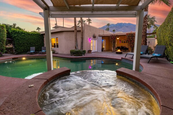 A hot tub with a waterfall effect, a pool, and a covered patio area with lounge chairs and a dining area.