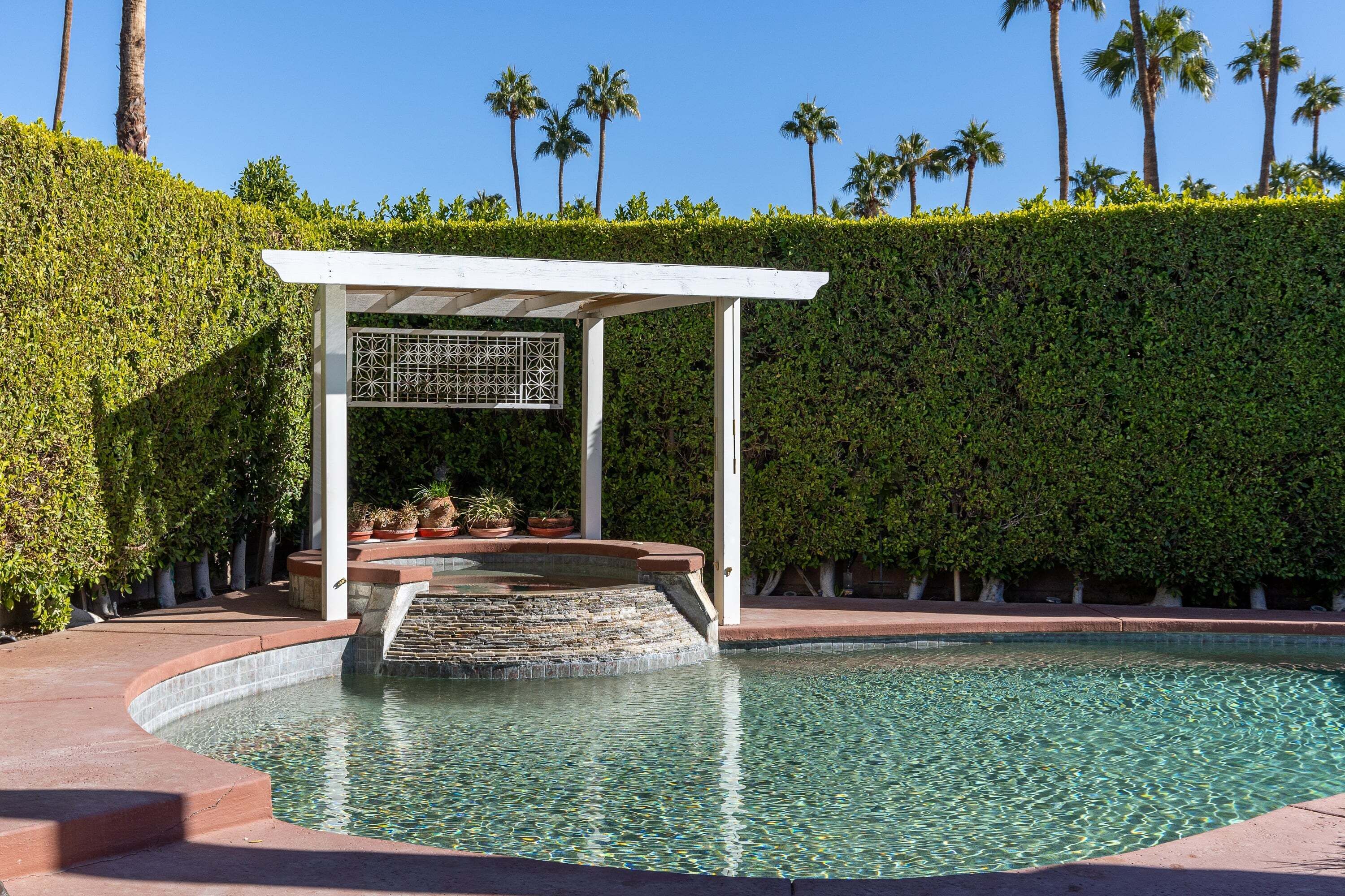 Ein Pool mit weißer Pergola, einem Steinelement und einer ordentlich geschnittenen Hecke.