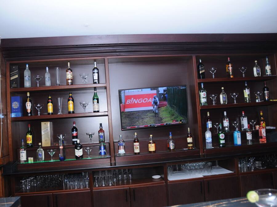 Zona de bar con estantes llenos de botellas de licor y vasos, un televisor mostrando un partido y una barra con copas y un trozo de limón.