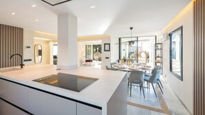 A modern kitchen with a central island, a dining area with a glass table, and a view of the outdoors. - Villa El Paraiso Marbella- 4 Bedrooms (5-Beds) (Benahavís)