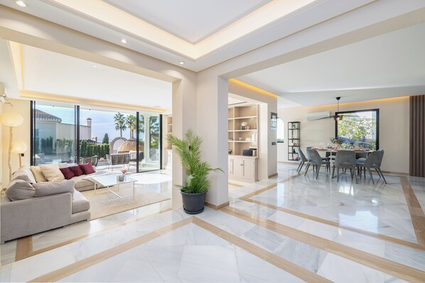 A spacious living area with a sofa, coffee table, and potted plant. There is a dining area with chairs and a table. The room has large windows and a view of the outside.