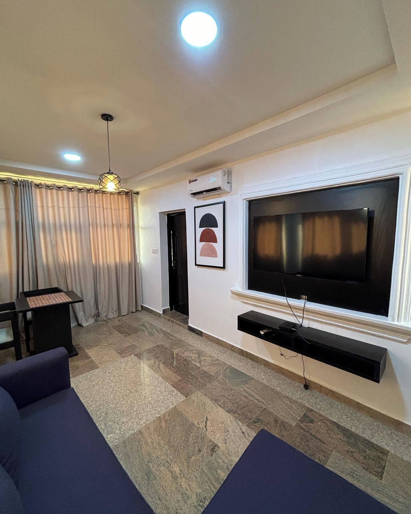 A living room with a large flat-screen TV mounted on the wall, a dark blue sofa, a small table, and a patterned floor.