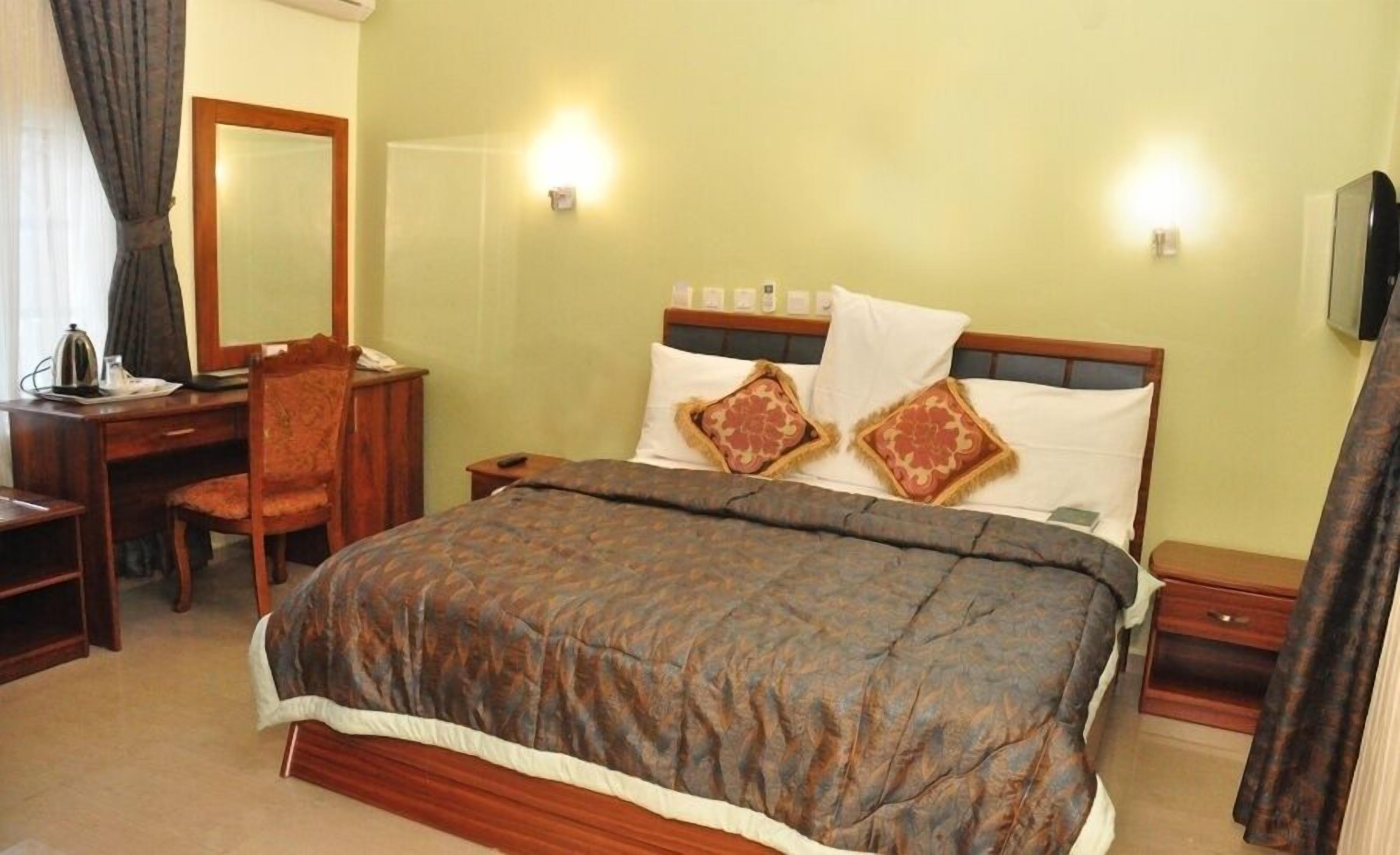 A hotel room with a bed, a desk, a chair, a bedside table, and a mirror.