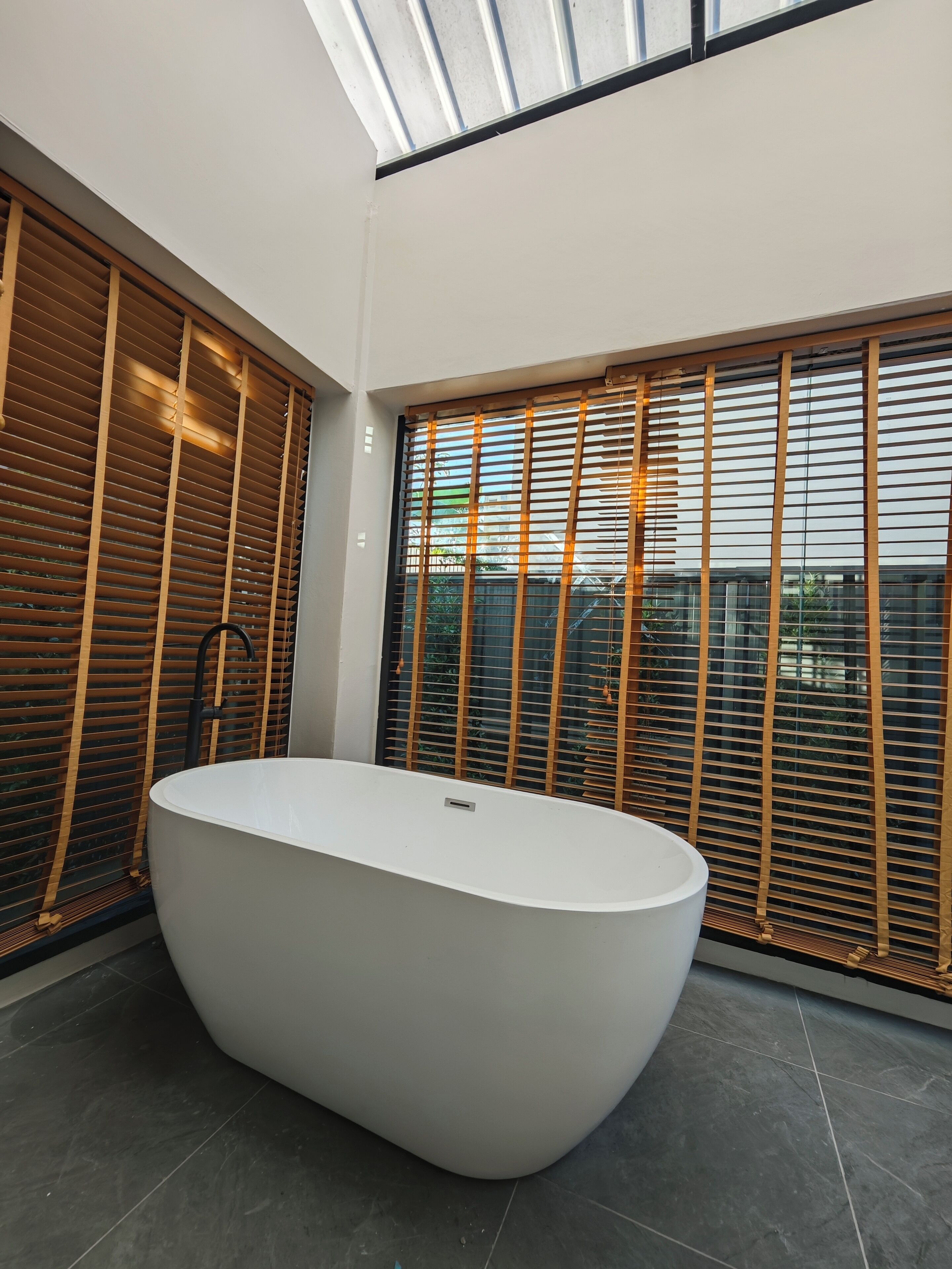A modern bathroom with a freestanding bathtub, large windows with blinds, and a skylight.