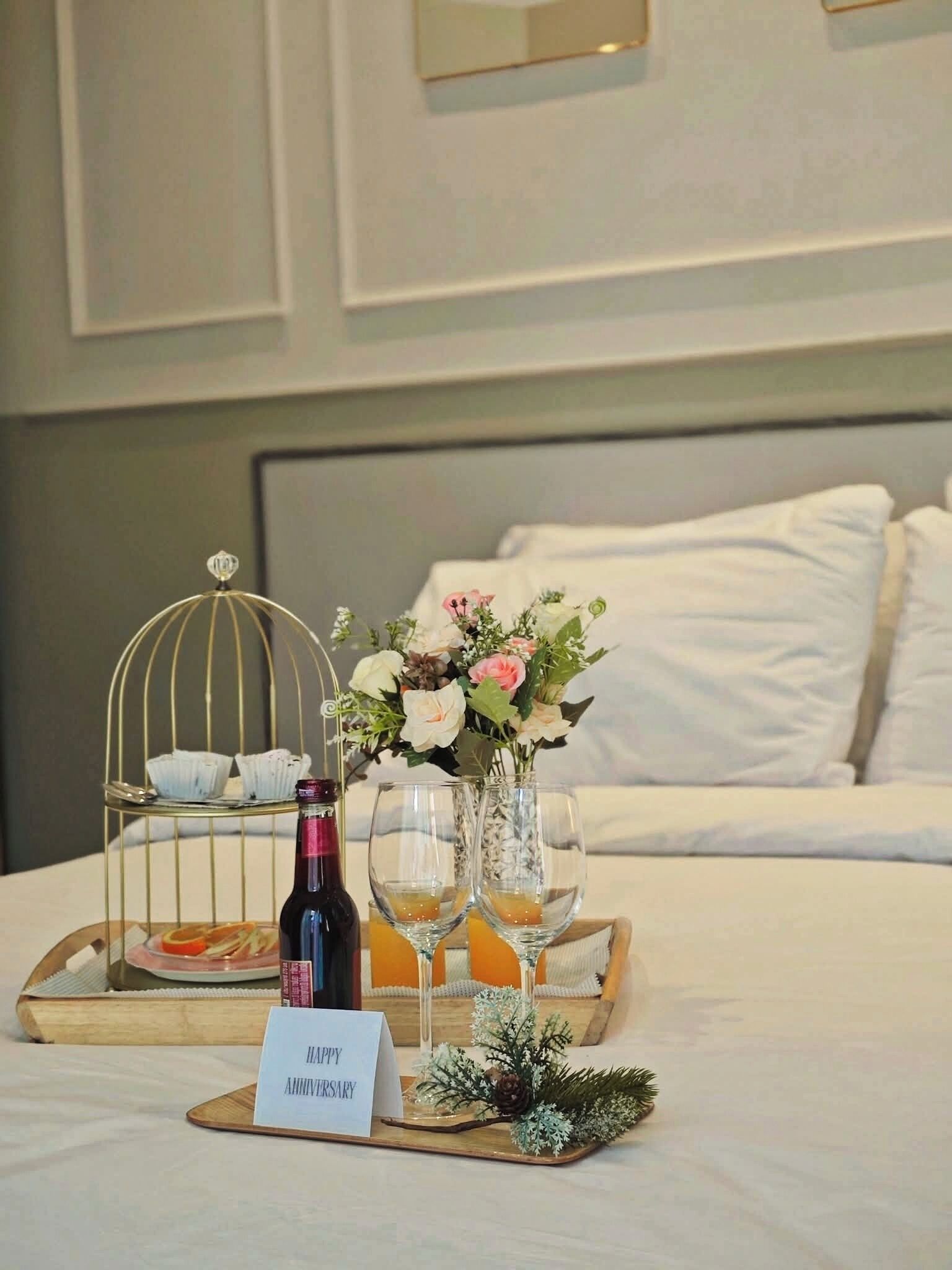 A neatly arranged breakfast tray on a bed with a 'HAPPY ANNIVERSARY' sign, a bottle of wine, and a floral arrangement.