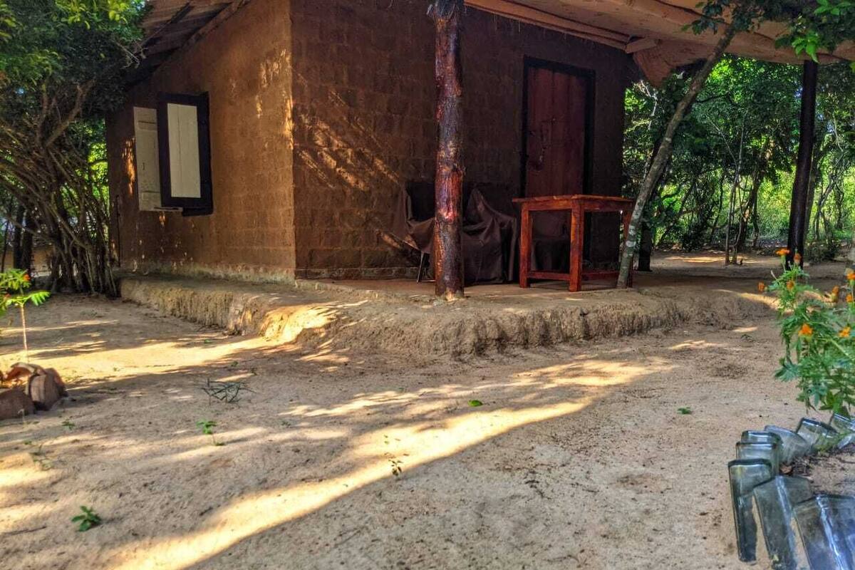 Una casa de madera rústica con suelo de tierra, una mesa de madera y una silla afuera.