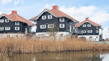A row of three black and white houses with red roofs, situated by a calm body of water with tall grasses in the foreground.