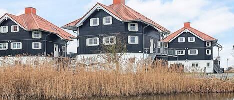 A row of three black and white houses with red roofs, situated by a calm body of water with tall grasses in the foreground.