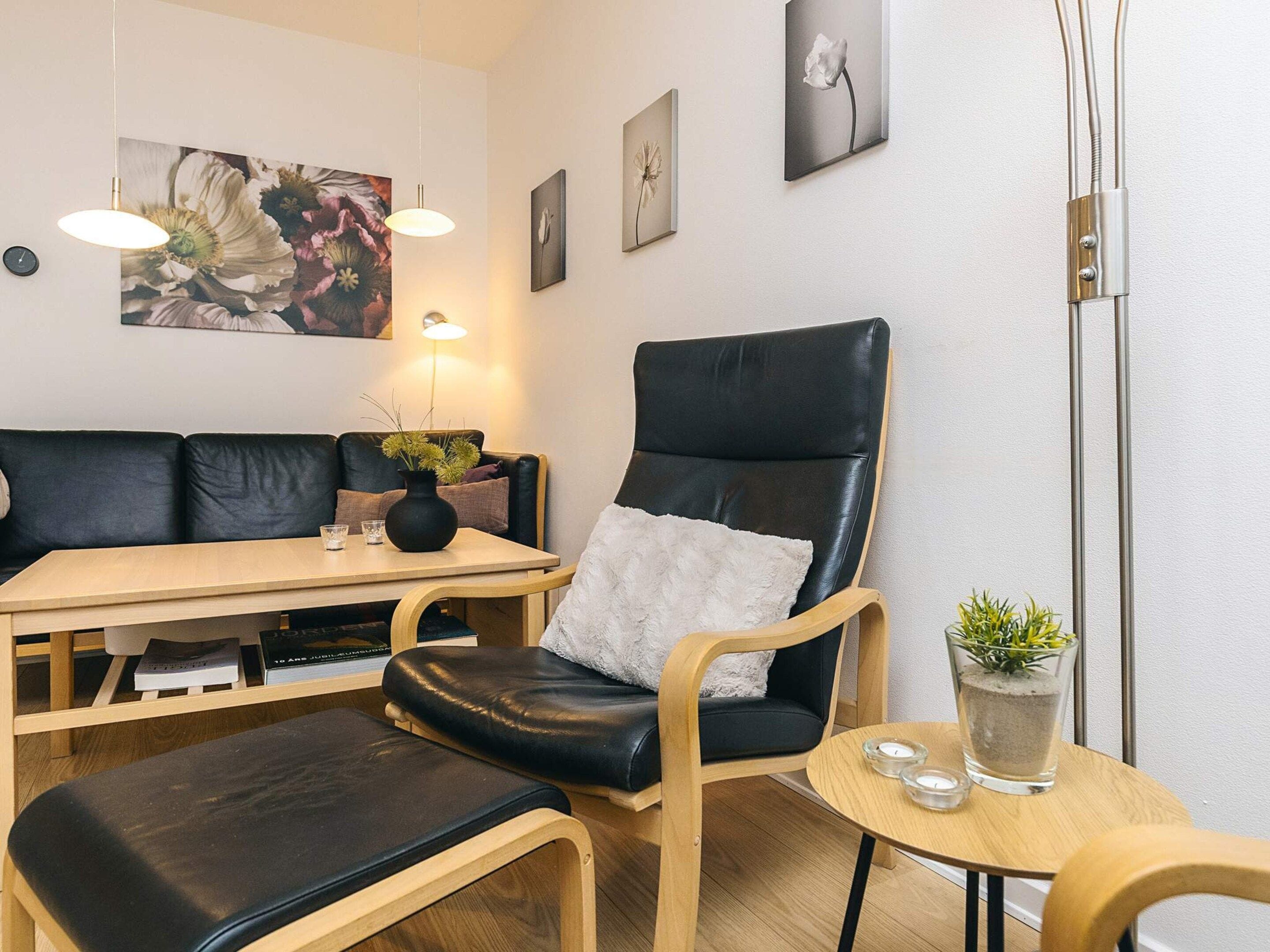 A modern living room with a black leather sofa, a wooden table, and a black leather armchair. There are framed pictures on the wall, a floor lamp, and a small table with a plant and candles.