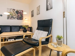 A modern living room with a black leather sofa, a wooden table, and a black leather armchair. There are framed pictures on the wall, a floor lamp, and a small table with a plant and candles.