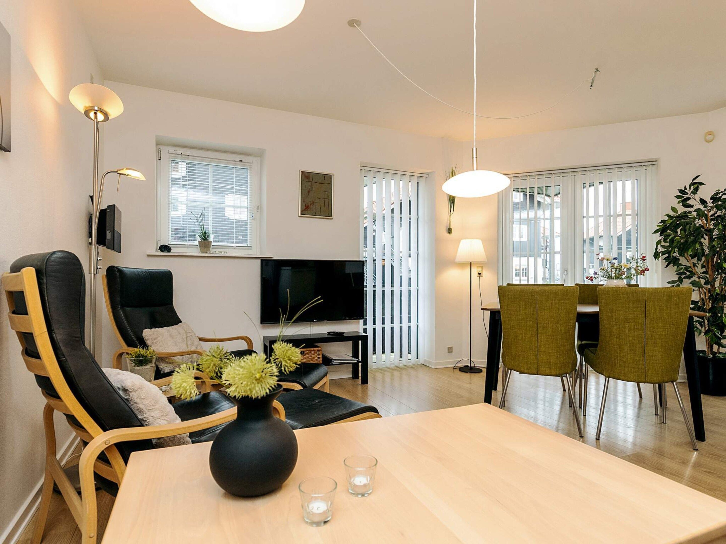 A modern living room with a TV, seating, a dining table, and a large window with blinds.