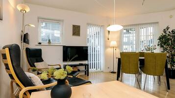 A modern living room with a TV, seating, a dining table, and a large window with blinds.