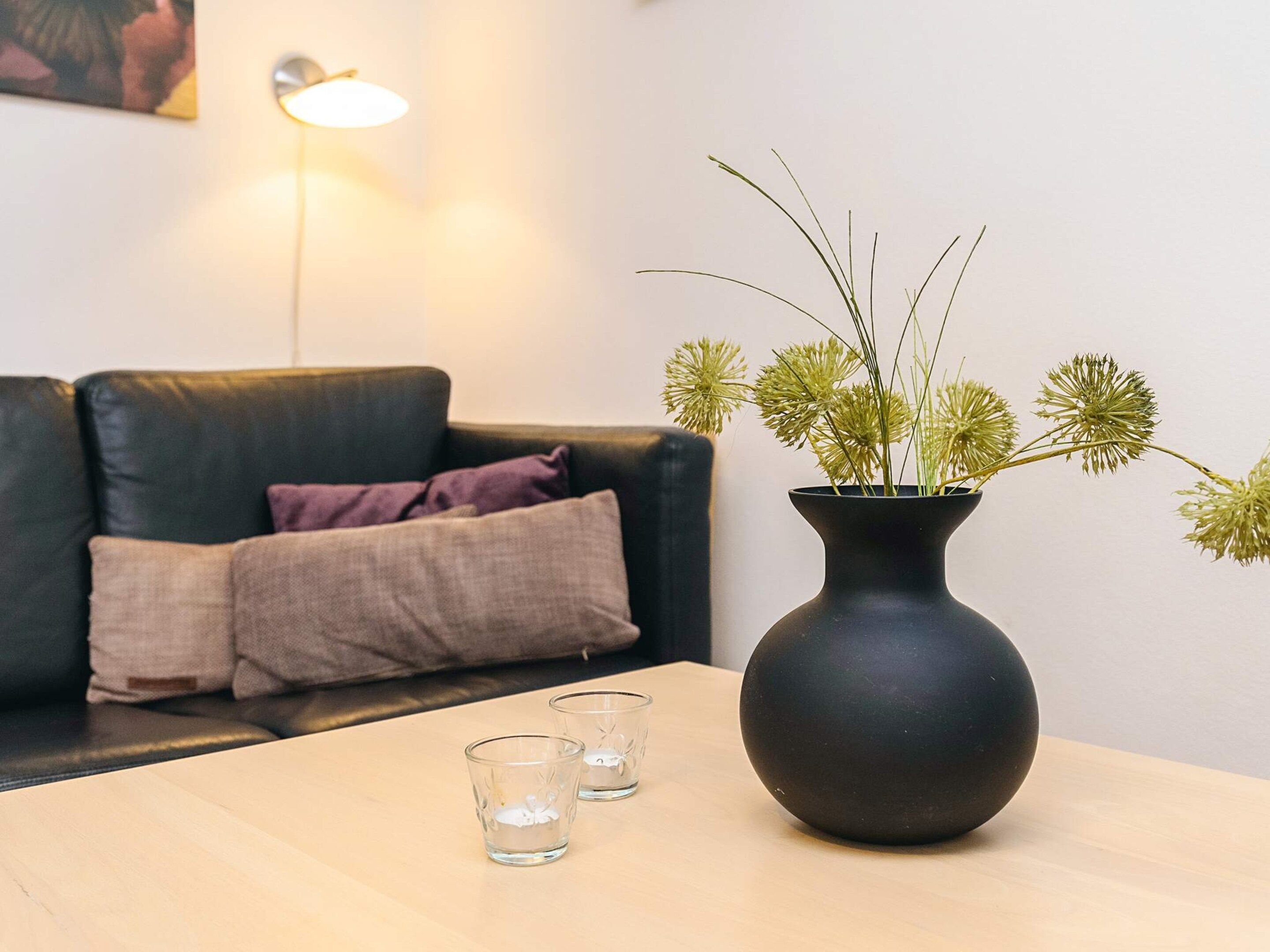 A living room with a dark leather sofa, a vase with dried flowers, and two glasses on a wooden table.