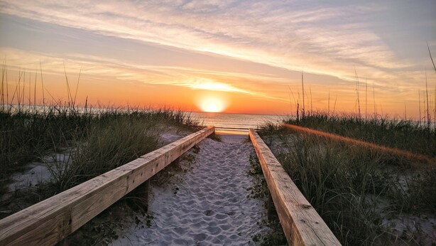 Ein Holzweg führt zum Strand, im Hintergrund ein Sonnenuntergang.