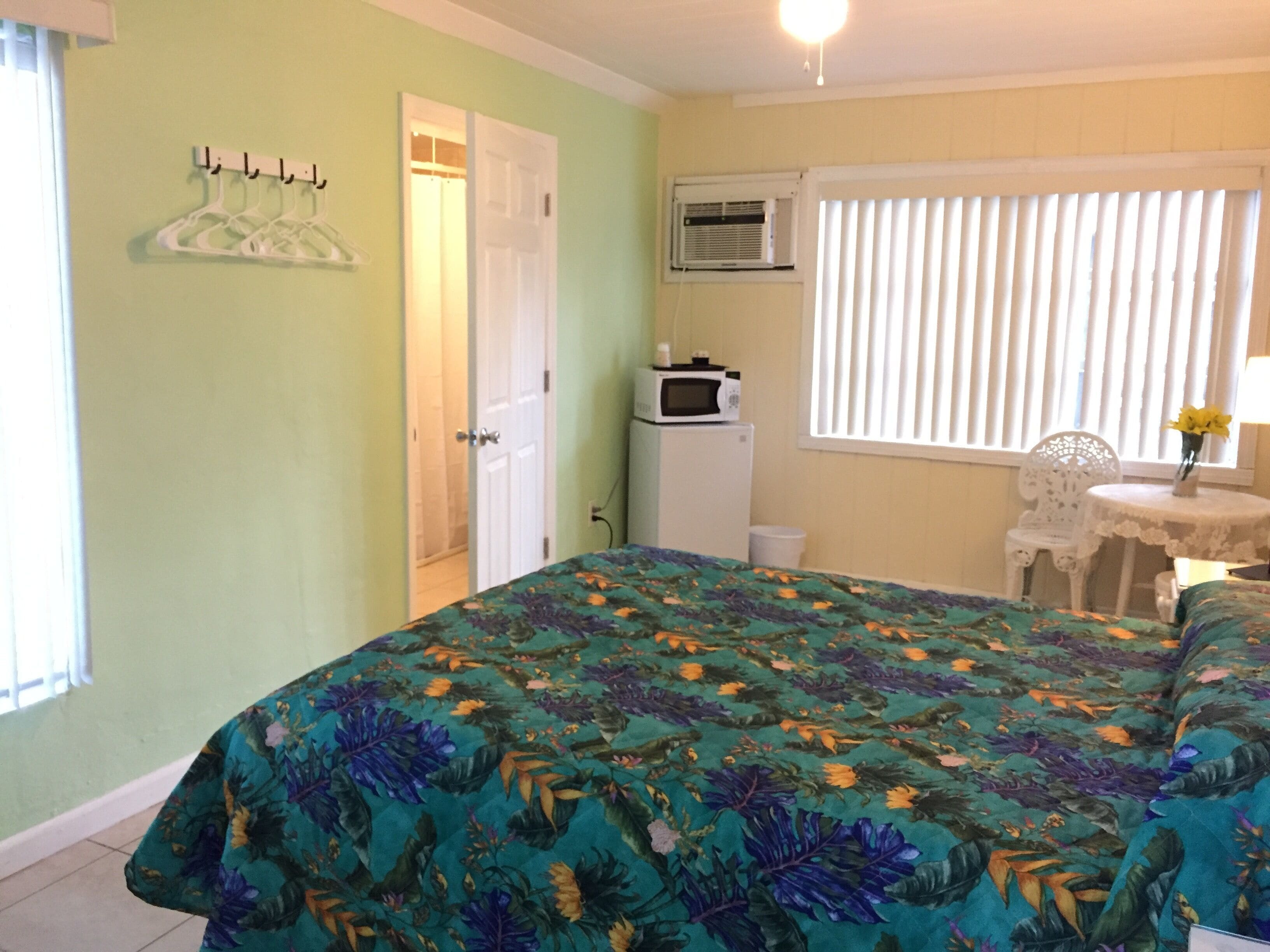 A bedroom with a bed, a small table with a chair, an air conditioning unit, and a door leading to another room.