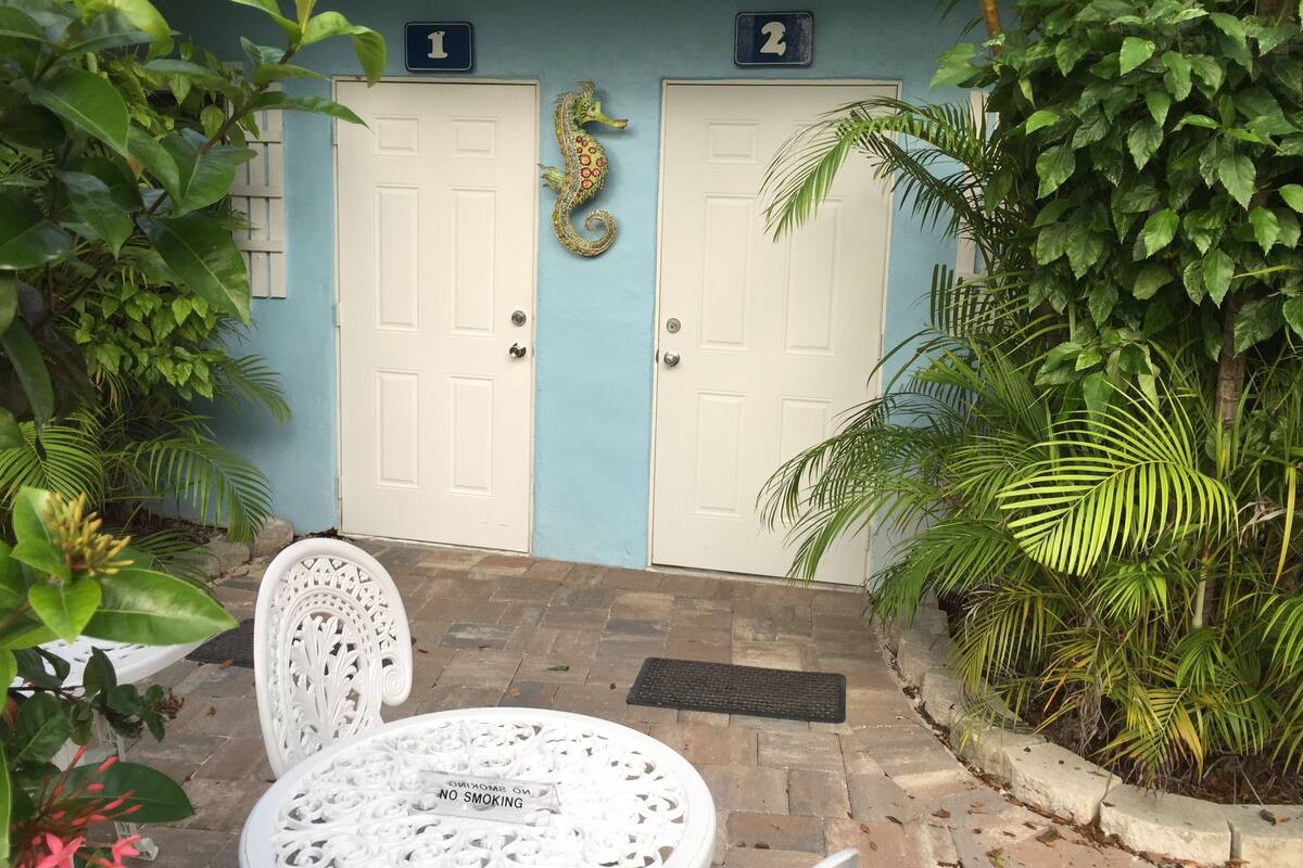 A two-room guest accommodation with numbered doors, a white chair, a round table, and a "No Smoking" sign.