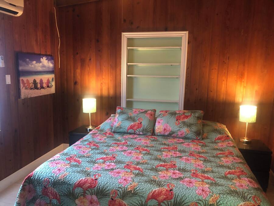 Et soveværelse med trævægge, en seng med blomstret sengetæppe, to sengeborde med lamper og et billede i ramme på væggen.