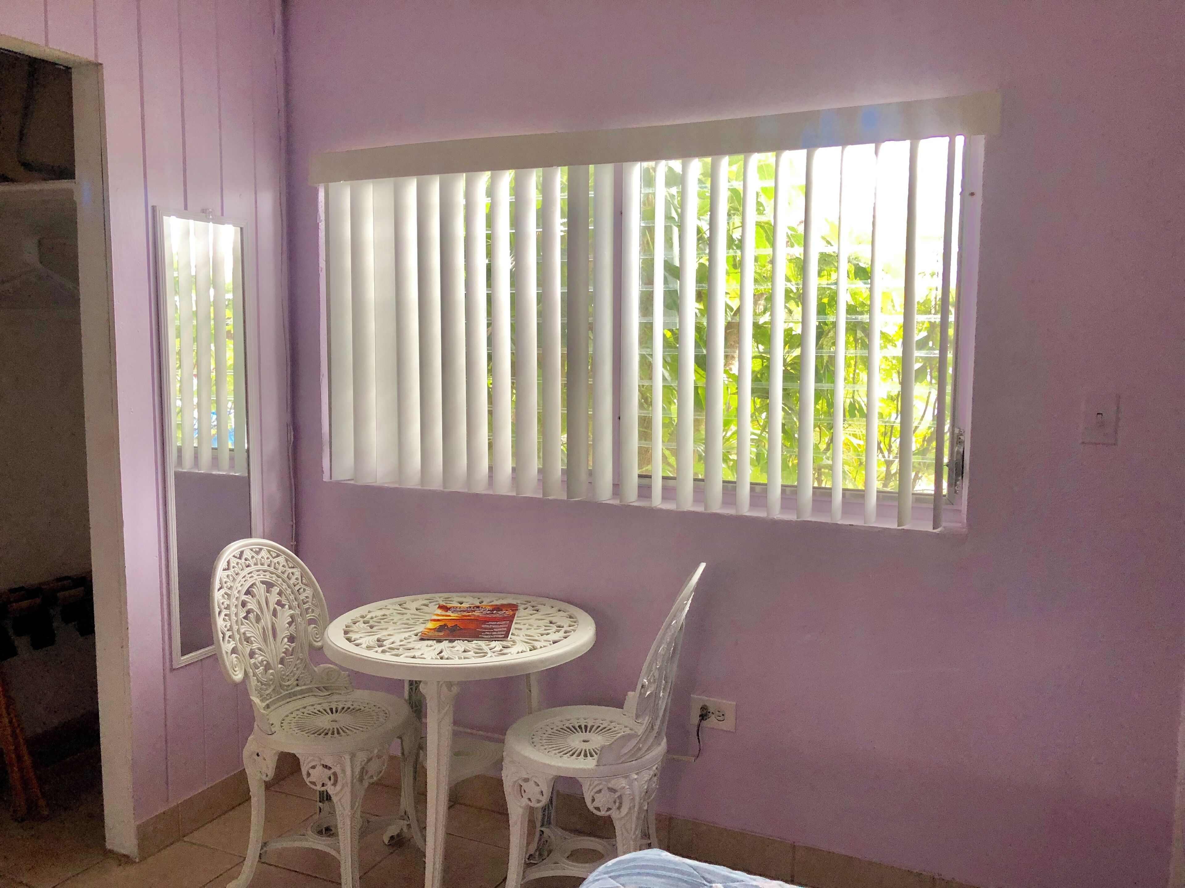A room with a purple wall, a window with vertical blinds, a round table with two chairs, and a mirror on the wall.