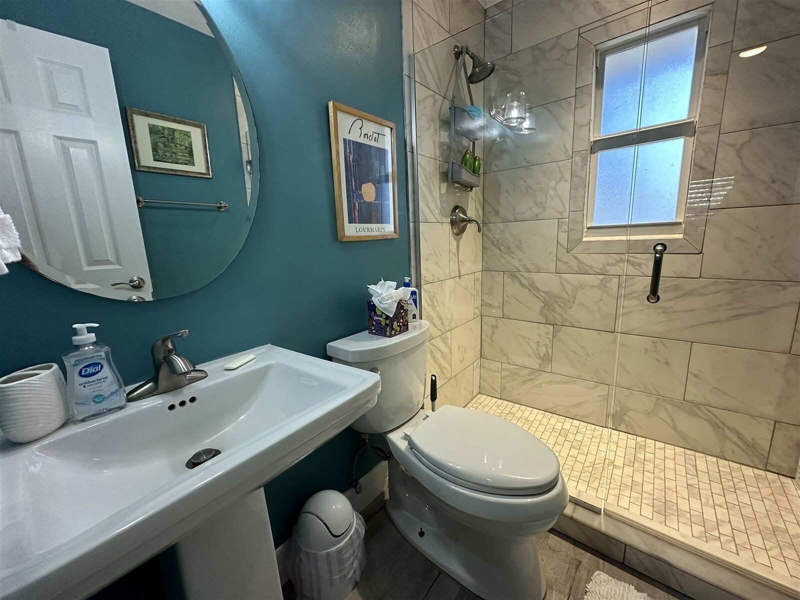 A bathroom with a white sink, toilet, and shower. There are framed pictures on the wall and a window above the shower.