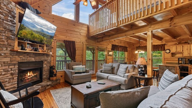 A cozy cabin interior with a stone fireplace, wooden furniture, and a balcony overlooking a forested area. - Triple Mountain View Stunning 4 BDR Cabin w/ Private Hot Tub, Game Room (Gatlinburg)