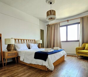Une chambre à coucher avec un lit en bois, deux tables de chevet avec des lampes, une fenêtre avec des rideaux et un fauteuil jaune.