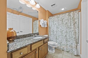Un baño con lavamanos de encimera de granito, tocador de madera, inodoro blanco y cortina de ducha con estampado floral.
