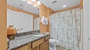 Un baño con lavamanos de encimera de granito, tocador de madera, inodoro blanco y cortina de ducha con estampado floral.