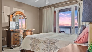 A hotel room with a bed, a dresser, a TV, and a balcony with a view of the sunset. - Legacy Tower II 609 | Stunning Coastal Views! (Gulfport)
