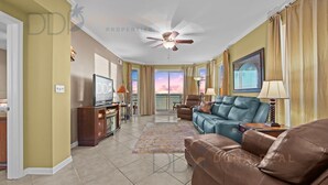 A spacious living room with a ceiling fan, a large window with curtains, a brown leather sofa, a blue sofa, and a television on a wooden stand. - Legacy Tower II 609 | Stunning Coastal Views! (Gulfport)