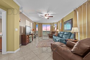 A spacious living room with a ceiling fan, a large window with curtains, a brown leather sofa, a blue sofa, and a television on a wooden stand. - Legacy Tower II 609 | Stunning Coastal Views! (Gulfport)