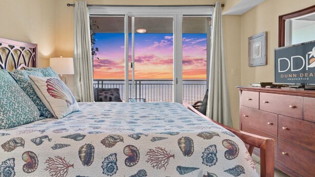 A bedroom with a bed, a dresser, a TV, and a balcony with a sunset view. - Legacy Tower II 907 | Deluxe Unit with Stunning Coastal Views! (Gulfport)