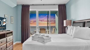 Habitación de hotel con una cama grande, una mesita de noche, una lámpara, un televisor y vistas a la puesta de sol por las ventanas.
