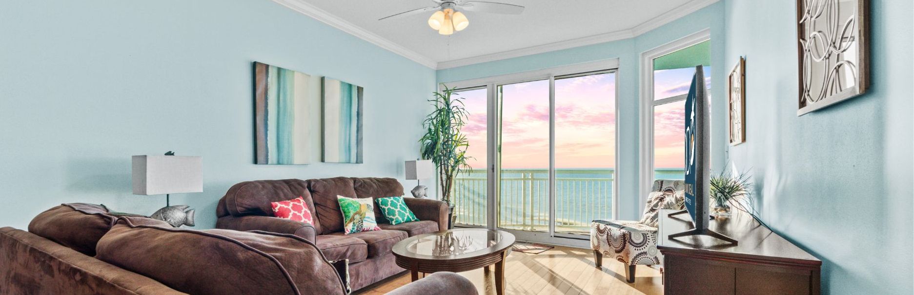 A living room with a brown sofa, colorful pillows, a glass coffee table, and a view of the ocean through large sliding doors.