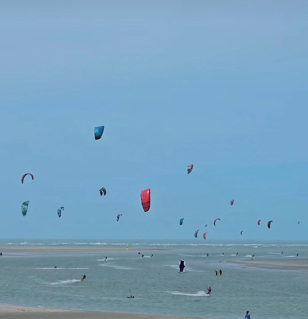 A beach with multiple people engaged in kite surfing, and several kites visible in the sky.