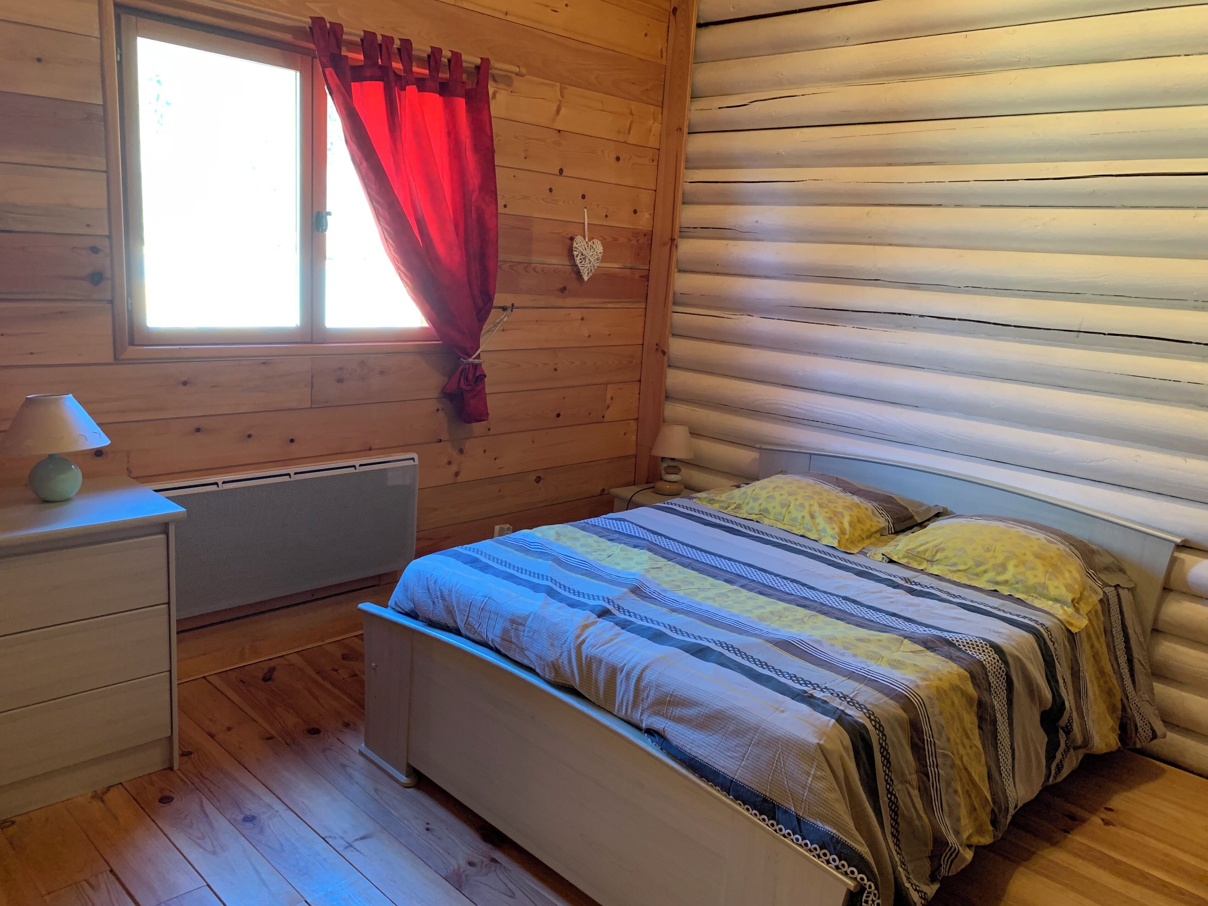 Ein gemütliches Blockhauszimmer mit Holzvertäfelung, einem Bett mit gemusterten Textilien, einem Nachttisch mit Lampe und einem Fenster mit roten Vorhängen.