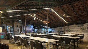 Un vaste espace intérieur aménagé en salle à manger, avec de nombreuses tables et chaises, un plafond haut avec des poutres apparentes et un mur orné d’affiches informatives.